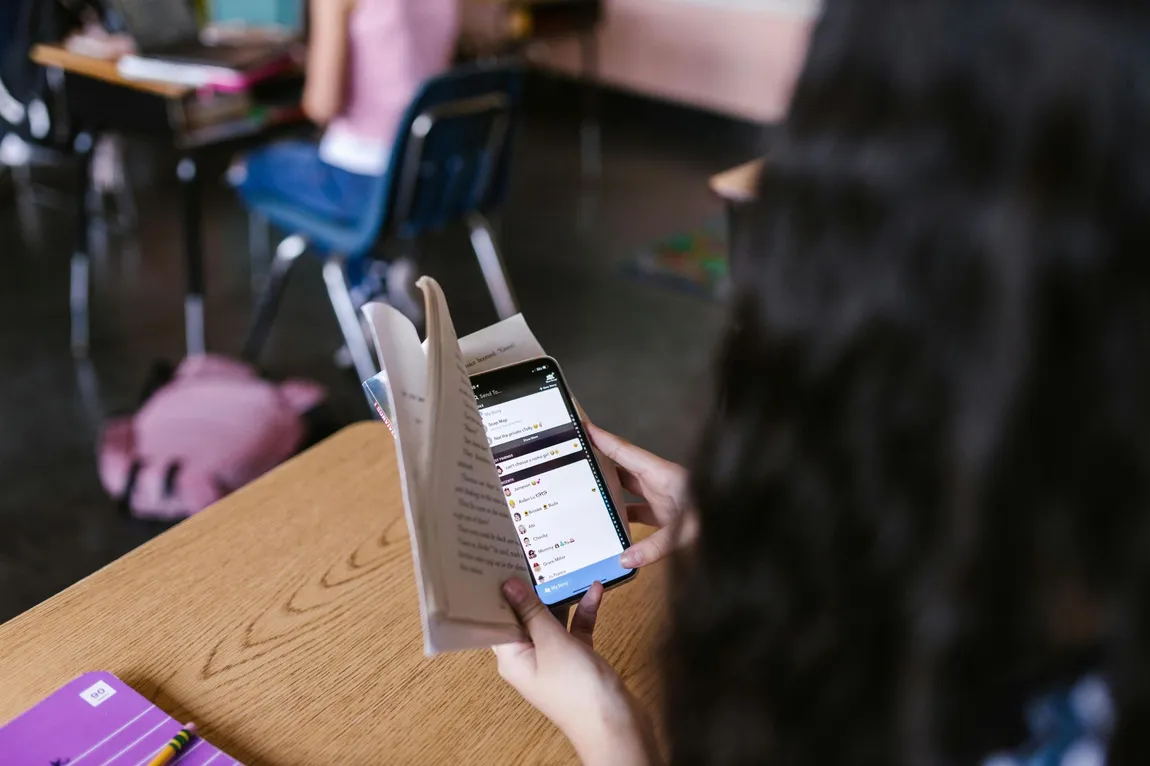 Uno studente al banco di scuola usa silenziosamente il telefono, tenendolo basso dietro un libro aperto